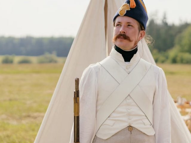 A historical reenactor in Napoleonic military uniform standing by a tent in open countryside.