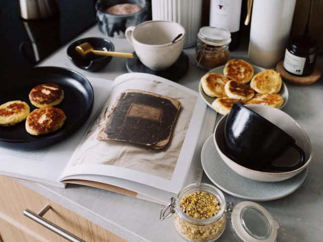 High angle of cookbook and cooked fritters on plates placed on table near utensils and ingredients in cozy kitchen at home